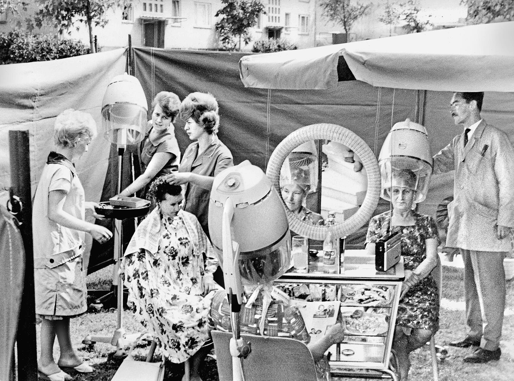 Wer schön sein will, muss leiden, umso mehr, wenn ein Friseurbesuch in der Hitze des Sommers ansteht. Ein erfindungsreicher Figaro aus Fürth verlegte deshalb kurzerhand im Jahre 1963 seinen Salon ins Freie und verschönerte mit Schere, Kamm und Lockenwicklern seine Kundinnen „open-air“. Abgeschirmt von neugierigen Blicken freuten sich die Damen mit frisch eingedrehten Haaren bei Radiomusik und entspannender Lektüre, wenn sie neben der heißen Fön-Luft auch eine kühle Brise unter die Trockenhaube bekamen.