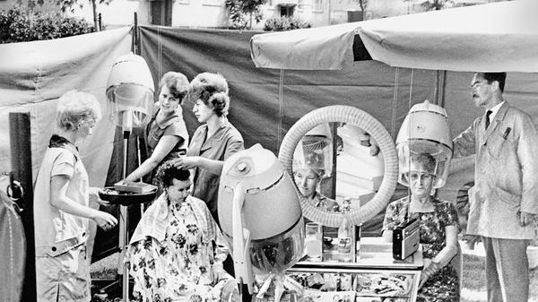 Wer schön sein will, muss leiden, umso mehr, wenn ein Friseurbesuch in der Hitze des Sommers ansteht. Ein erfindungsreicher Figaro aus Fürth verlegte deshalb kurzerhand im Jahre 1963 seinen Salon ins Freie und verschönerte mit Schere, Kamm und Lockenwicklern seine Kundinnen „open-air“. Abgeschirmt von neugierigen Blicken freuten sich die Damen mit frisch eingedrehten Haaren bei Radiomusik und entspannender Lektüre, wenn sie neben der heißen Fön-Luft auch eine kühle Brise unter die Trockenhaube bekamen.