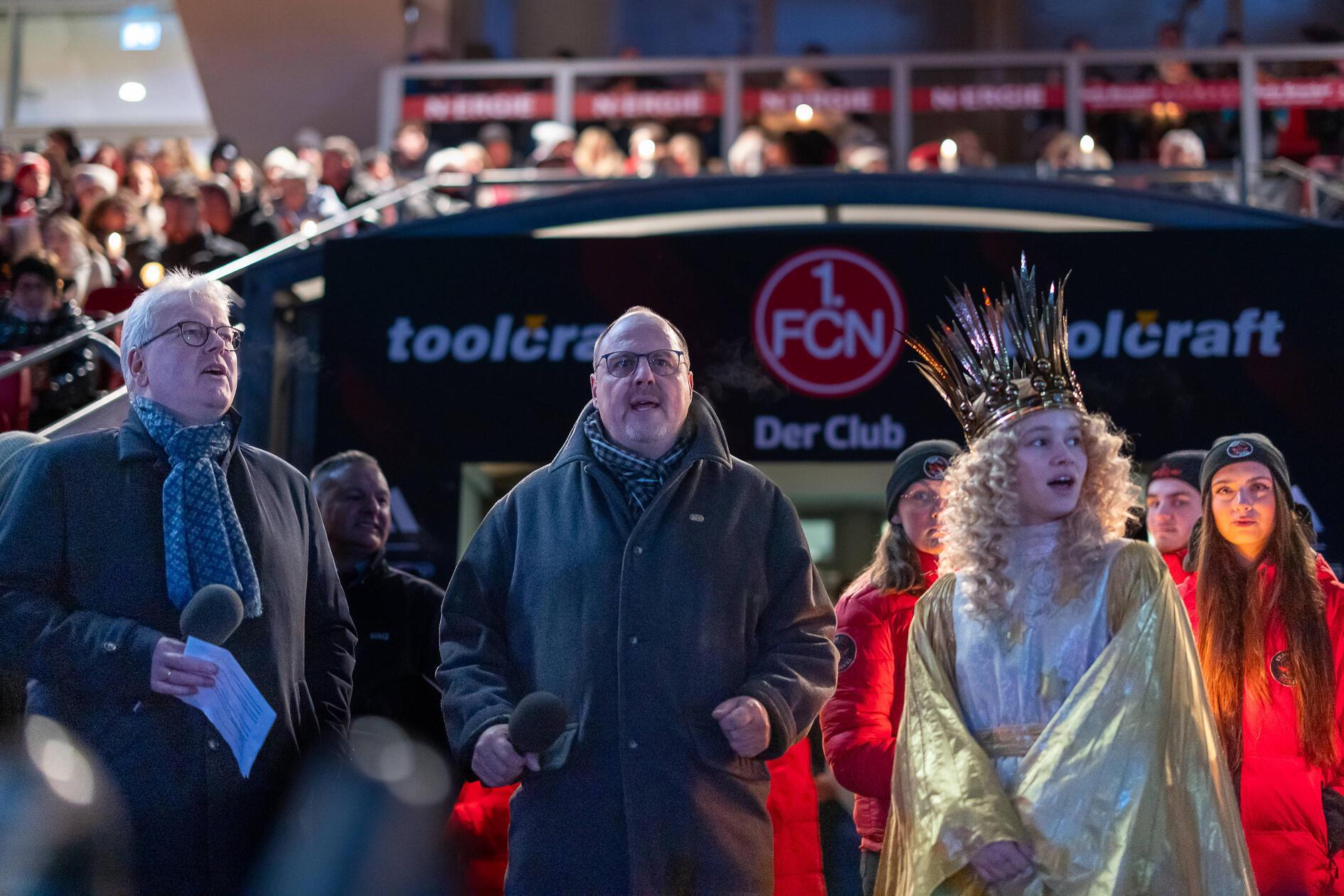 Alle singen gemeinsam: Links Dr. Matthias Everding, Vorstandsvorsitzender der Sparkasse Nürnberg, in der Mitte Bürgermeister Christian Vogel von der SPD, rechts das festlich gekleidete Nürnberger Christkind Nora Falk. Vogel sagte, es sei einzigartig, kurz vor Weihnachten gemeinschaftlich mit Gesang in die Weihnachtszeit zu starten. In seinen Augen vereint es „jedes Jahr aufs Neue in einer außergewöhnlichen Atmosphäre im Max-Morlock-Stadion Gemeinschaft und die Freude daran, miteinander zu singen und gleichzeitig Gutes füreinander zu tun.“