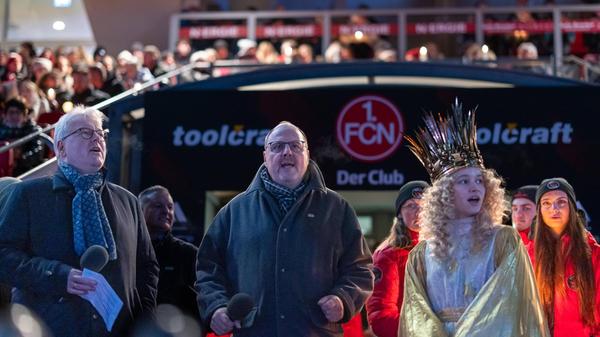 Alle singen gemeinsam: Links Dr. Matthias Everding, Vorstandsvorsitzender der Sparkasse Nürnberg, in der Mitte Bürgermeister Christian Vogel von der SPD, rechts das festlich gekleidete Nürnberger Christkind Nora Falk. Vogel sagte, es sei einzigartig, kurz vor Weihnachten gemeinschaftlich mit Gesang in die Weihnachtszeit zu starten. In seinen Augen vereint es „jedes Jahr aufs Neue in einer außergewöhnlichen Atmosphäre im Max-Morlock-Stadion Gemeinschaft und die Freude daran, miteinander zu singen und gleichzeitig Gutes füreinander zu tun.“