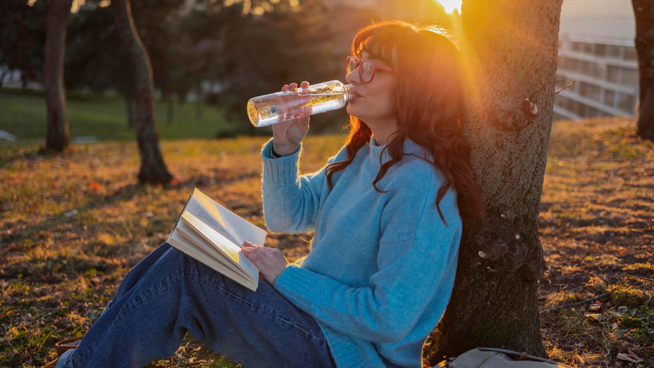Woman drinking water and sitting with book near tr
