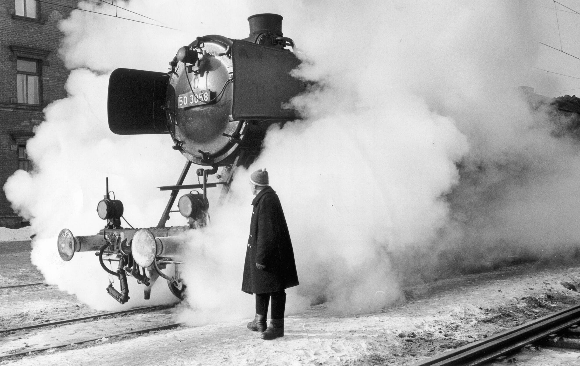 Von Dampf umhüllt, so lichtete unsere Fotografin im Februar 1963 diese archaisch anmutende alte Lokomotive ab, die in dieser dramatischen Einstellung an „The General“ aus dem gleichnamigen Buster Keaton – Film erinnert. In Ermangelung eines vorne an der Lok angebrachten Kuhfängers hat der Herr im Vordergrund seinerzeit jedoch glücklicherweise darauf verzichtet, die Stunts des Stummfilmstars nachzuahmen.