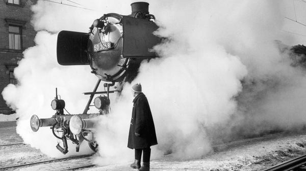 Von Dampf umhüllt, so lichtete unsere Fotografin im Februar 1963 diese archaisch anmutende alte Lokomotive ab, die in dieser dramatischen Einstellung an „The General“ aus dem gleichnamigen Buster Keaton – Film erinnert. In Ermangelung eines vorne an der Lok angebrachten Kuhfängers hat der Herr im Vordergrund seinerzeit jedoch glücklicherweise darauf verzichtet, die Stunts des Stummfilmstars nachzuahmen.