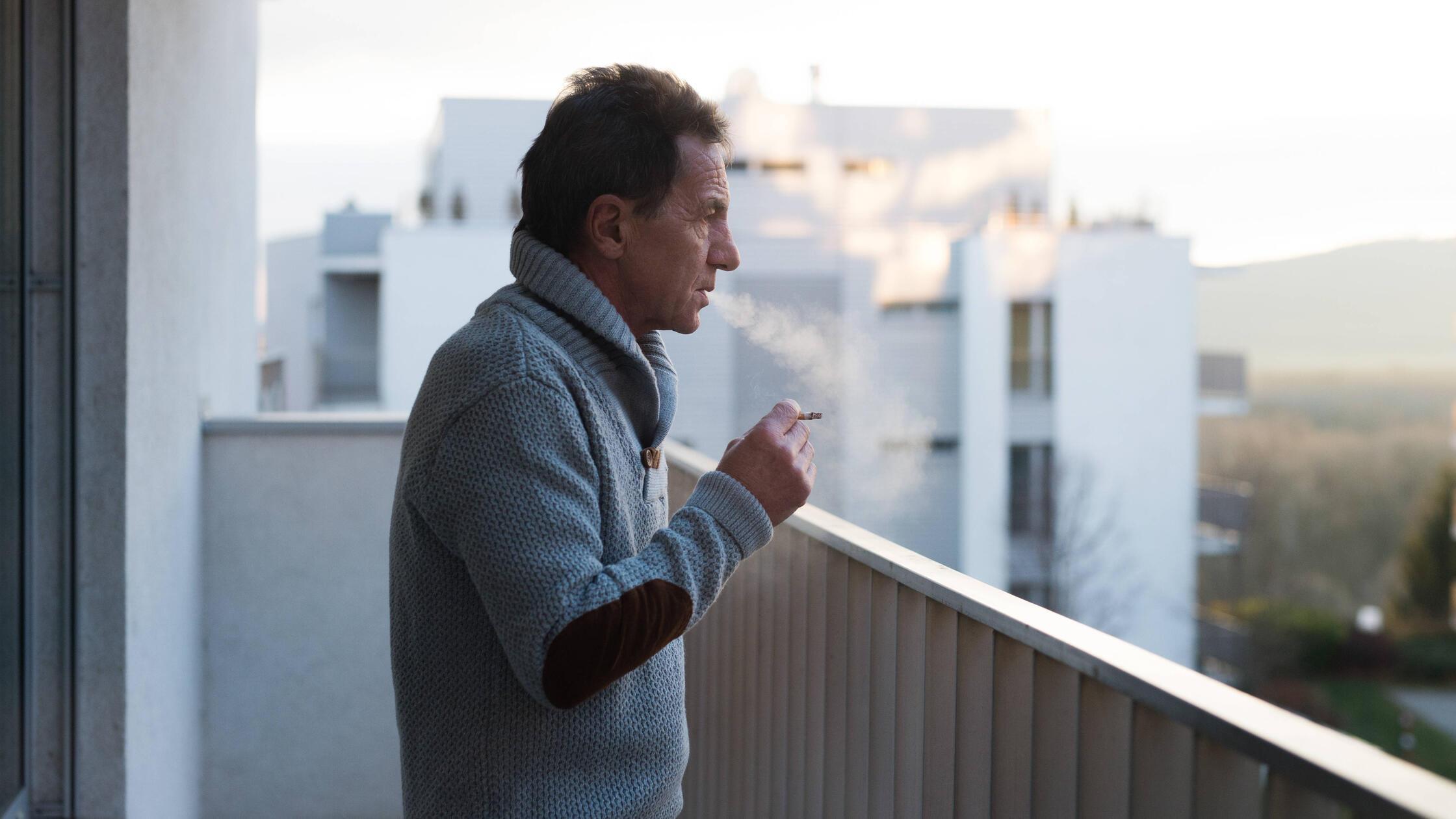 Serious senior man standing on balcony, smoking a 