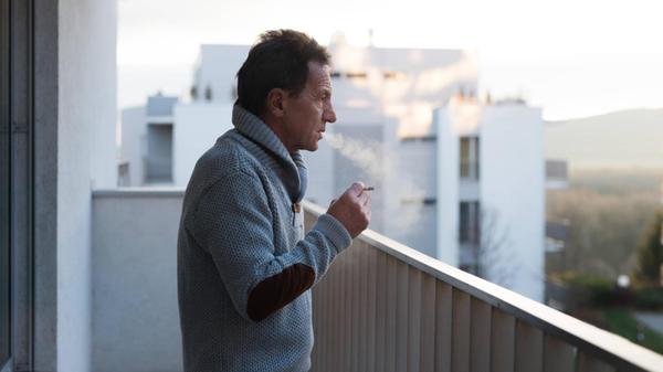 Serious senior man standing on balcony, smoking a