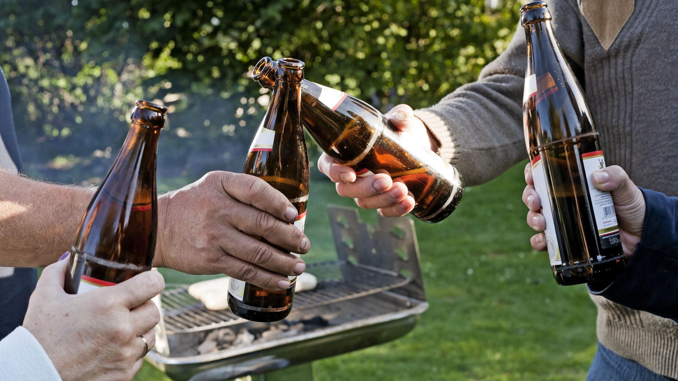 Vier Leute stoßen draußen vor einem Grill mit Bier