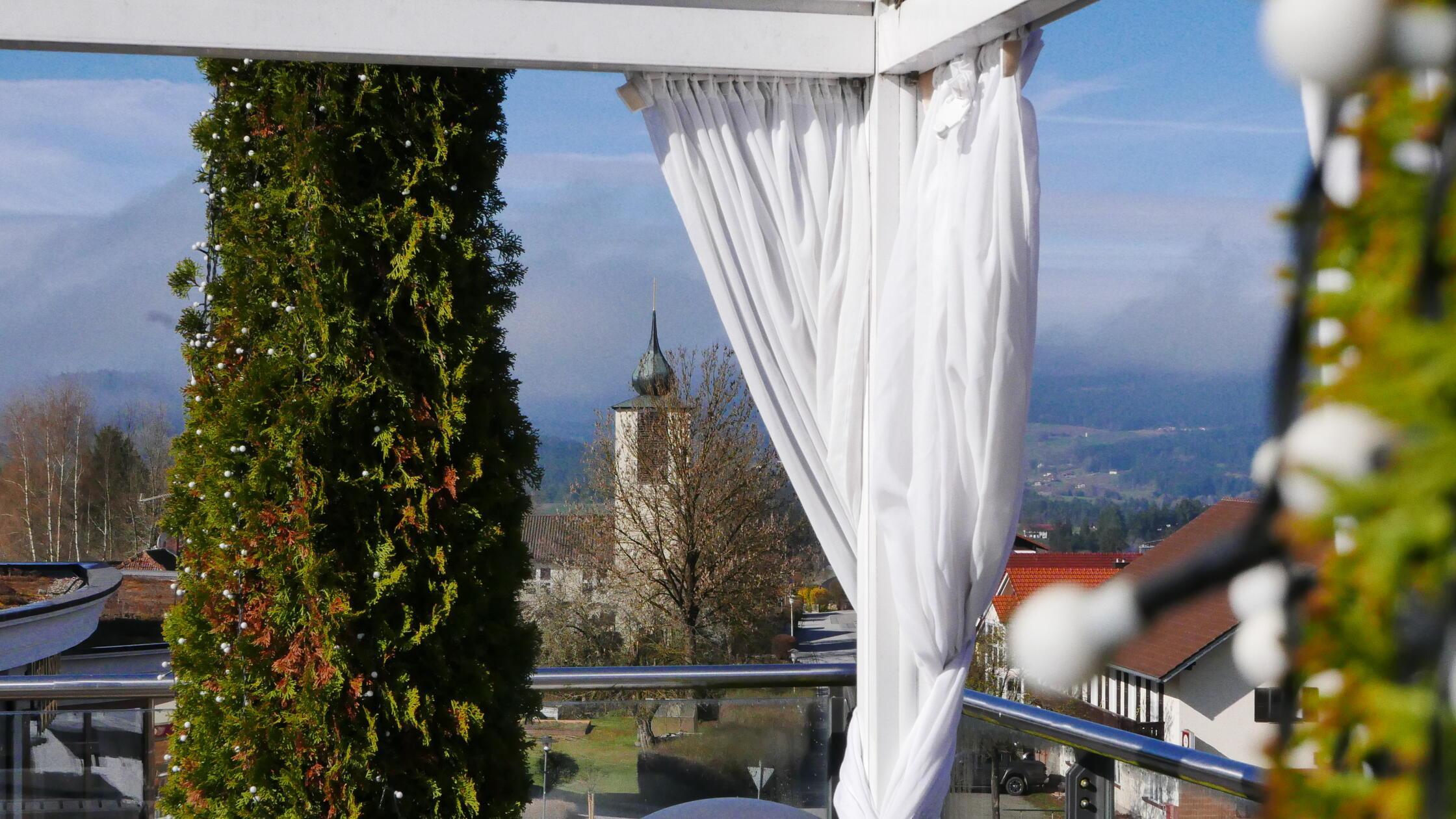 Blick durch weiße Vorhänge auf die hölzerne Terrasse, dahinter der Kirchturm von Kaikenried.