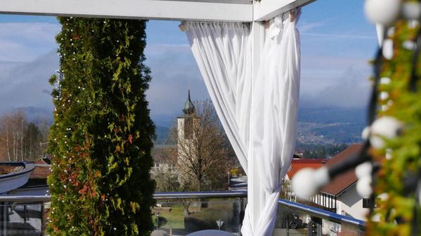 Blick durch weiße Vorhänge auf die hölzerne Terrasse, dahinter der Kirchturm von Kaikenried.
