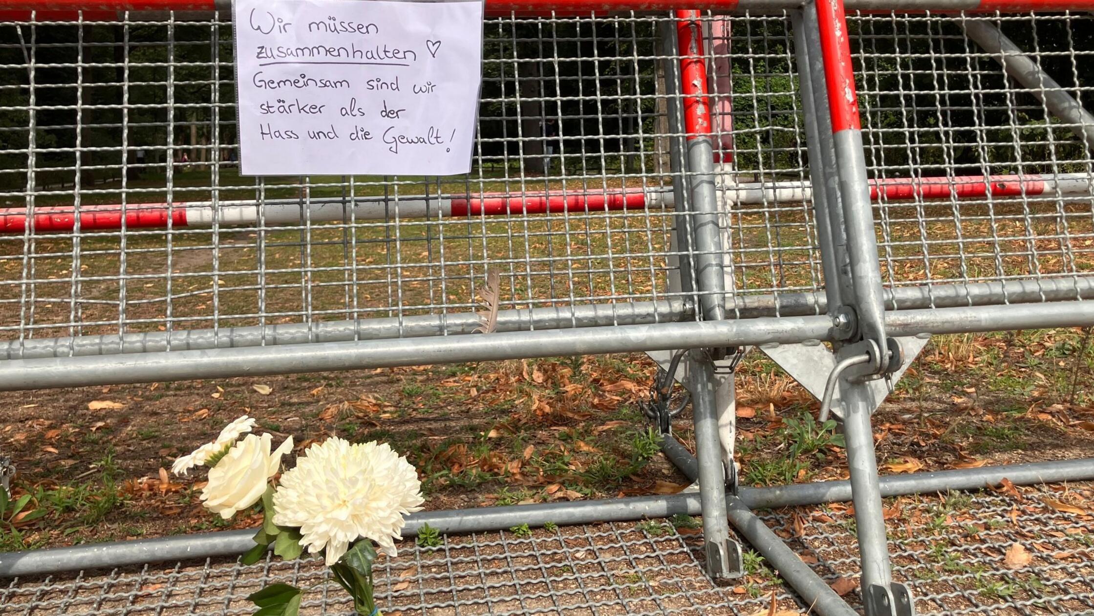 An einem Zaun, an dem Blumen liegen, hängt ein Zet
