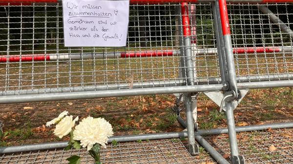 An einem Zaun, an dem Blumen liegen, hängt ein Zet