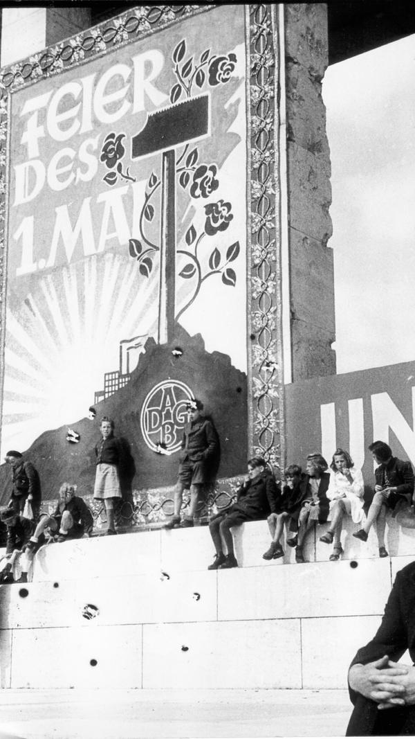 unter einem Maiplakat, das einen blühenden Hammer als Zeichen friedlicher Arbeit und eine aufgehende Sonne als Symbol des neuen Anfangs zeigt. Die NN schrieben: "Der schönste Gedanke aber war, daß diese Menschen alle freiwillig gekommen waren, um für den neuen Sinn der Arbeit, für friedlichen Wiederaufbau und für internationale Verständigung zu demonstrieren."