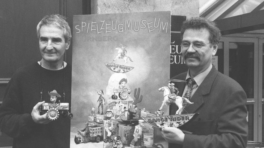 Fotograf Lajos Keresztes (l.) und Dr. Helmut Schwarz halten das Plakat von 2001 in der Hand. Es zeigt mechanisches Spielzeug, das zwischen 1930 und 1950 in New York gefertigt wurde.