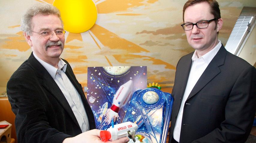 Der Leiter des Spielzeugmusems, Dr. Helmut Schwarz (l.), und Grafiker Udo Bernstein widmen das Jahresplakat 2009 des Spielzeugmuseums der Erde-Mond-Bahn der Firma Gebrüder Einfalt.