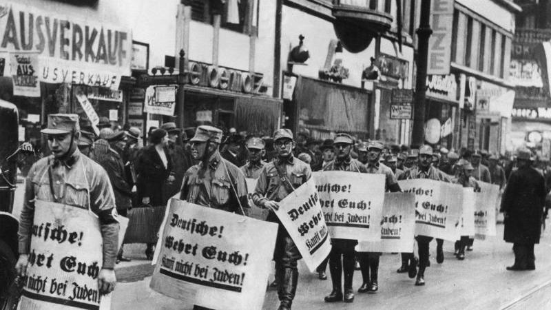 Männer der SA im Jahr 1938 bei einem Hetzmarsch gegen die jüdische Bevölkerung Deutschlands. Die zur Schau getragenen Plakate tragen die Aufschrift "Deutsche wehrt Euch! Kauft nicht bei den Juden!".
