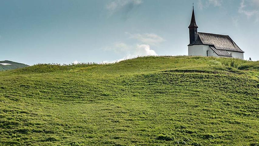Eine Kirche und eine Almkuh im Warth. Die Landschaft wirkt auf Touristen wie gemalt.
