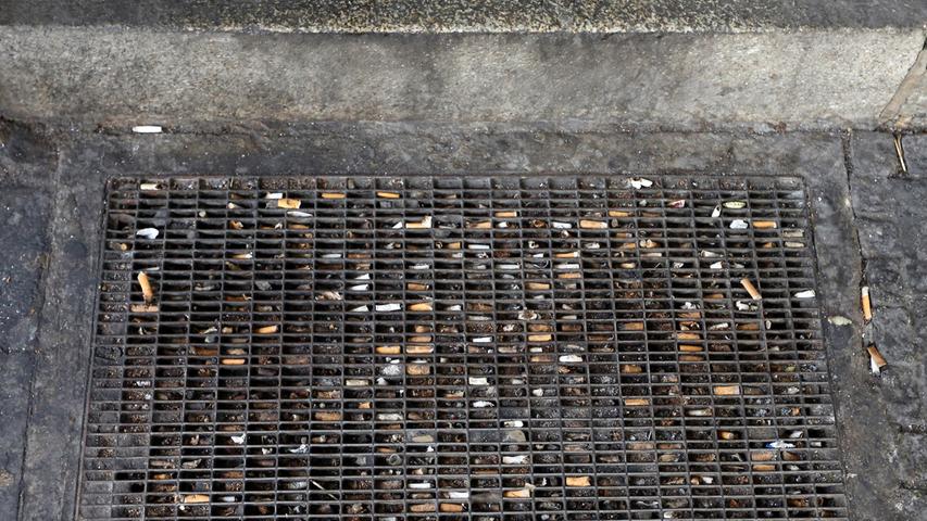 In diesem Gitter, welches im von einer schwarzen Schmutzschicht überzogenen Boden eingelassen ist, stecken unzählige vergilbte Zigarettenstummel.