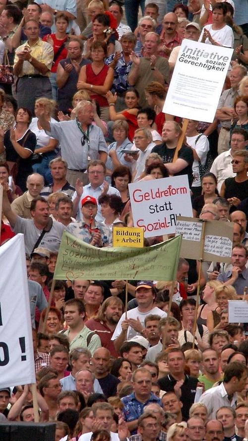 Als Reaktion auf das umstrittene Hartz-Konzept kommt es in Deutschland zu Massenprotesten. Mit Transparenten stehen Demonstranten am Montag, 16. August 2004, auf dem Leipziger Simsonplatz. Unter dem Motto "Weg mit Hartz IV und Agenda 2010 - Für Arbeit, Gerechtigkeit und Solidarität" protestierten mehr als 25 000 Menschen gegen die geplanten Einschnitte im Sozialsystem durch die Reformen von Hartz IV und der Agenda 2010.