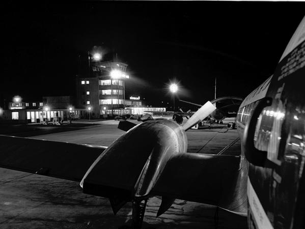 Der Flughafen mit seinem charakteristischen Turm im Glanz der Scheinwerfer und Lampen. Auf dem Vorfeld stehen eine Passagiermaschine vom Typ Vickers Viscount (hinten), die eben aus Düsseldorf angekommen ist, und ein Postflugzeug. Gerade die zuschlagfreie Nachtpost hat das Aufkommen um mehr als das Zwanzigfache steigen lassen. Ein einmaliger Rekord. Der Flughafen mit seinem charakteristischen Turm im Glanz der Scheinwerfer und Lampen. Auf dem Vorfeld stehen eine Passagiermaschine vom Typ Vickers Viscount (hinten), die eben aus Düsseldorf angekommen ist, und ein Postflugzeug. Gerade die zuschlagfreie Nachtpost hat das Aufkommen um mehr als das Zwanzigfache steigen lassen. Ein einmaliger Rekord.