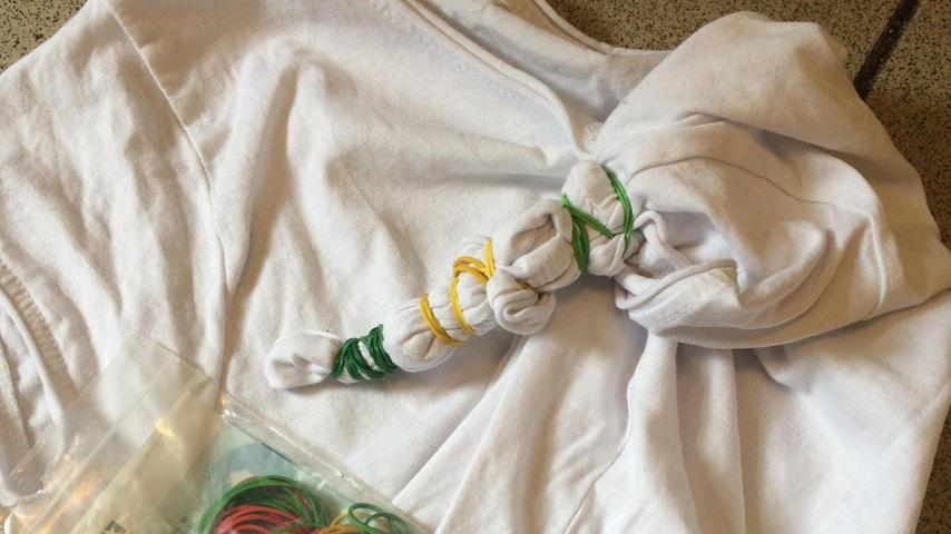 Wir haben ein altes weißes T-Shirt genommen, Gummi­bänder, Batikfarbe, einen alten Topf, einen Löffel zum Umrühren und Gummihandschuhe. Die gewünschte Stelle oder die gewünschten Stellen mit Gummibändern fest abbinden, mehre­re Gummibänder ergeben mehrere Ringe