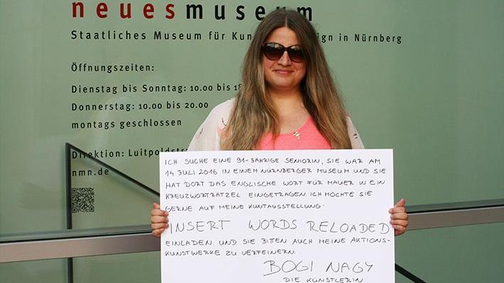 Die Nürnberger Künstlerin Bogi Nagy hält das Plakat hoch, mit dem sie die Kreuzworträtsel-Oma aus dem Neuen Museum sucht.