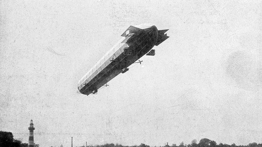 Diese Postkarte zeigt den Flug eines Zeppelins über den Dutzendteich im Jahr 1909. Links im Bild ist der eigens für die Landesausstellung 1906 errichtete Leuchtturm zu sehen.
