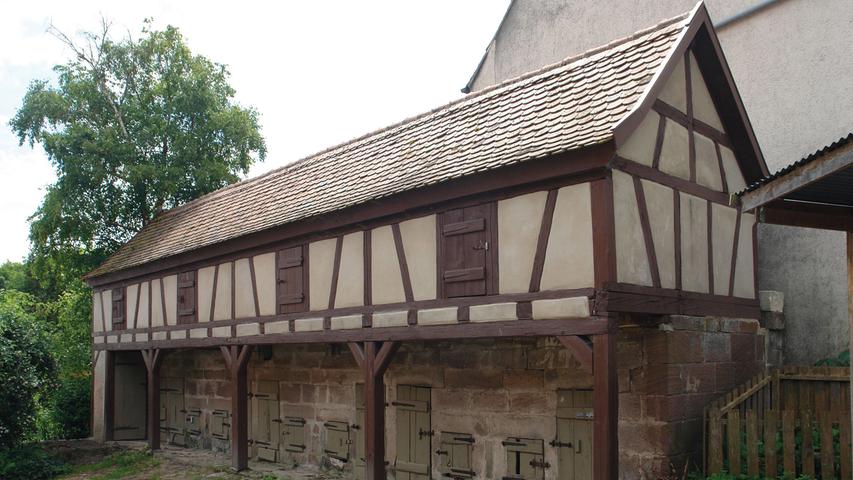 Auch beim Schweinestall musste das Fachwerk repariert werden. Hier wurden außerdem der Ortgang und die Stirnbretter erneuert; ebenso überarbeitete man die Dachfläche, bevor die Fassade des Stalles in Beige-Brauntönen getüncht wurde. Den Abschluss der Maßnahme bildet die Toreinfahrt. Ihre Rechteckpfosten mit Kugelaufsätzen haben ein neues Fundament bekommen und werden auf diese Weise geschickt präsentiert. Der naturbelassene Holzzaun komplettiert die Umzäunung. Angesichts der häufigen Leerstände historischer Bauten auf dem Land verdient diese Maßnahme Anerkennung. Gerade ungenutzte Wirtschaftsgebäude wie der Schweinestall werden viel zu häufig sich selbst überlassen. Wie sehr ihre Instandsetzung einem ganzen Ort zu Gute kommt, zeigt der Fall Obernbibert hervorragend.