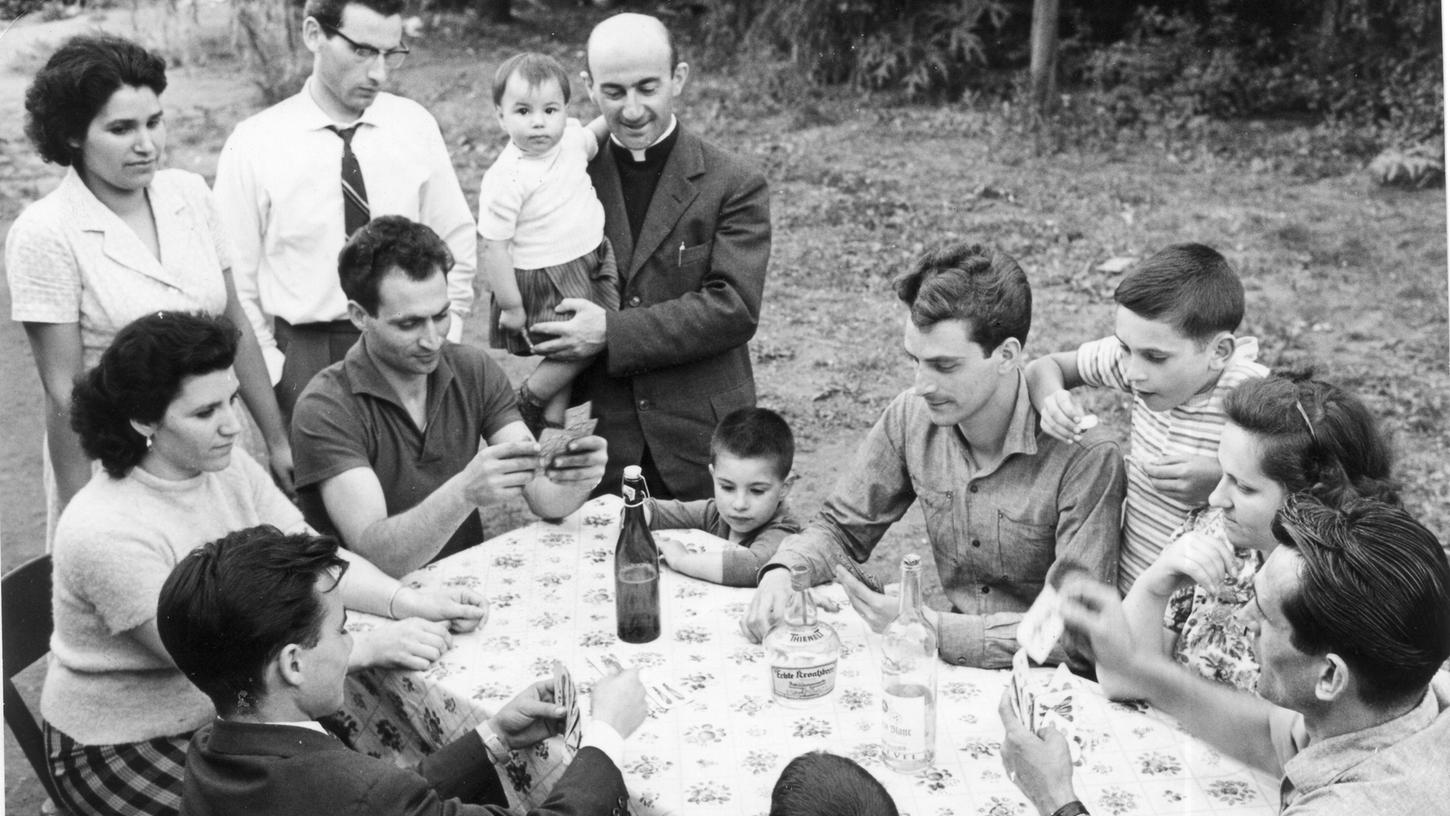 Der italienische Pfarrer Don Giuseppe besucht zusammen mit dem Sozialbetreuer Mario Ruzzu (li neben dem Pfarrer) einige Landsleute, die bereits ihre Familien hierhergebracht haben. Mit Bierflaschen und Kartenspiel wirken sie schon sehr akklimatisiert.