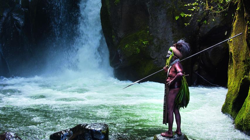 Ein Stammes-Chef posiert in Papua-Neuguinea auf einem Felsen vor einem Wasserfall.