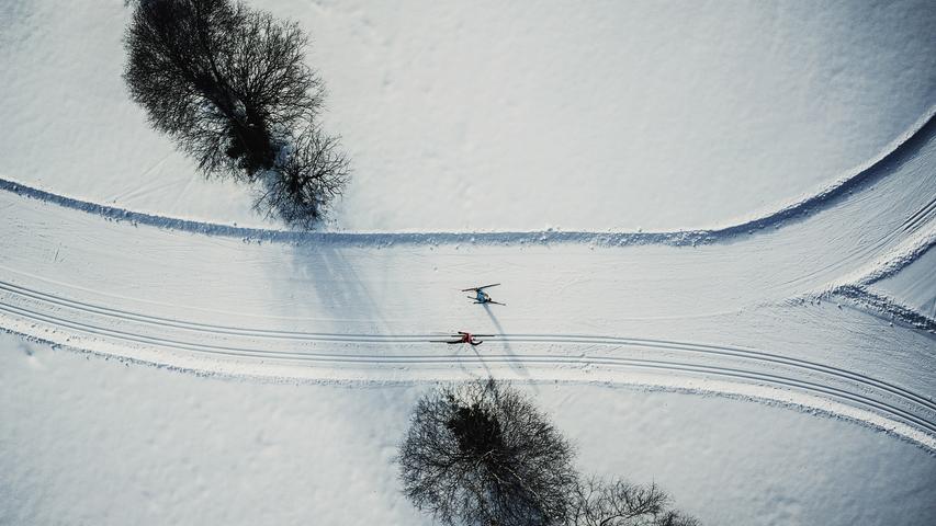 Bäume in einem Winterwunderland, Skilangläufer, die wie bunte Farbkleckse auf einer weißen Fläche wirken — die Drohne zeigt die Welt von oben.
