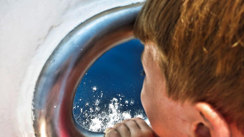 Gespannt schaut ein kleiner Junge durch das Loch für das Tau hinaus aufs blaue Wasser.