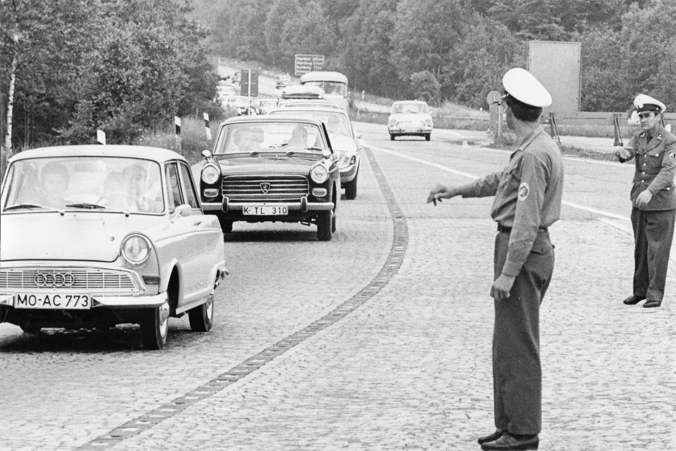 An einem Samstagvormittag im Juli 1965 waren diese Autos auf dem "Nürnberger Kreuz" der Autobahnen 3 und 9 Richtung München unterwegs. Man beachte das Kopfsteinpflaster der Ausfahrtsfahrbahn. Die Polizeibeamten waren vor Ort, um den Verkehrsfluss zu überwachen. Ihre Signale an die Autofahrer, zügiger zu fahren, seien oft vergeblich gewesen, ist überliefert. Ein DKW F12 führt auf diesem Bild die Karawane an, gefolgt von einem Peugeot 404 und einem (Ford) Taunus 17M.