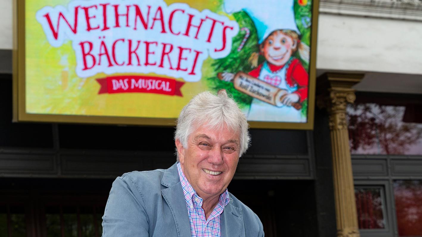 Rolf Zuckowski vor einem Plakat des 2019 in Nürnberg stattfindenden Kindermusicals "Die Weihnachtsbäckerei".