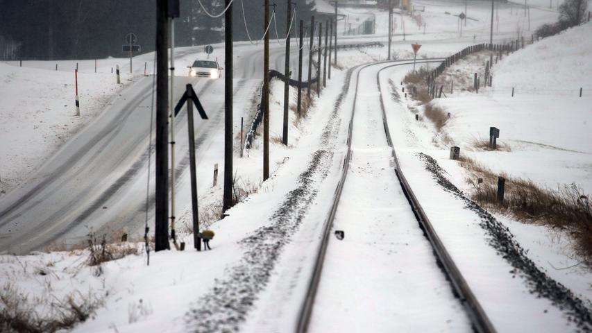 Eine weiße Schneedecke legt sich sowohl auf die Straße neben den Schienen, als auch über die Gleise der Bahnstrecke Füssen-Buchloe.