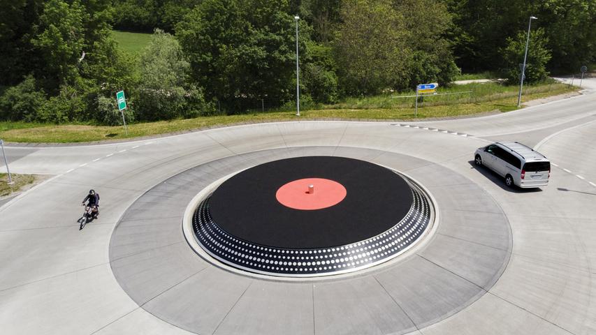 Ein Auto und eine Moped fahren in einem Kreisverkehr um einen Verkehrskreisel herum, der wie ein Plattenteller mit einer Vinyl-Platte aussieht. Das Kunstobjekt steht in Lyss im Kanton Bern.