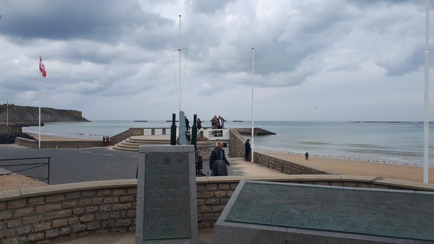 Am Strand von Arromanches erinnern Gedenk- und Informationstafeln an die Landung der alliierten Truppen im Juni 1944. Die Riesenblöcke im Wasser im Hintergrund sind Reste von damals angelegten künstlichen Befestigungen, um die Hafenzone vom offenen Meer abzugrenzen.