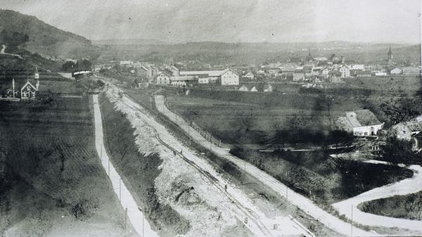Im Herbst 1869 fuhr der erste Zug von Treuchtlingen über Weißenburg nach Pleinfeld. Nach gut zwei Jahren Bauzeit war die Eröffnung der Strecke am 2. Oktober insbesondere für das bis dato unbedeutende Treuchtlingen ein Meilenstein, der die nächsten 100 Jahre Stadtgeschichte prägen sollte. Das Bild zeigt die provisorischen Gleise südlich der Stadt (links die Friedhofskapelle, rechts die Schmarrmühle) für den späteren Bau der Teilstrecke durchs Möhrenbachtal in Richtung Donauwörth. Der Bahndamm der Trasse Ingolstadt-Treuchtlingen-Weißenburg-Pleinfeld ist in der Bildmitte als dunkler Balken zu erkennen.