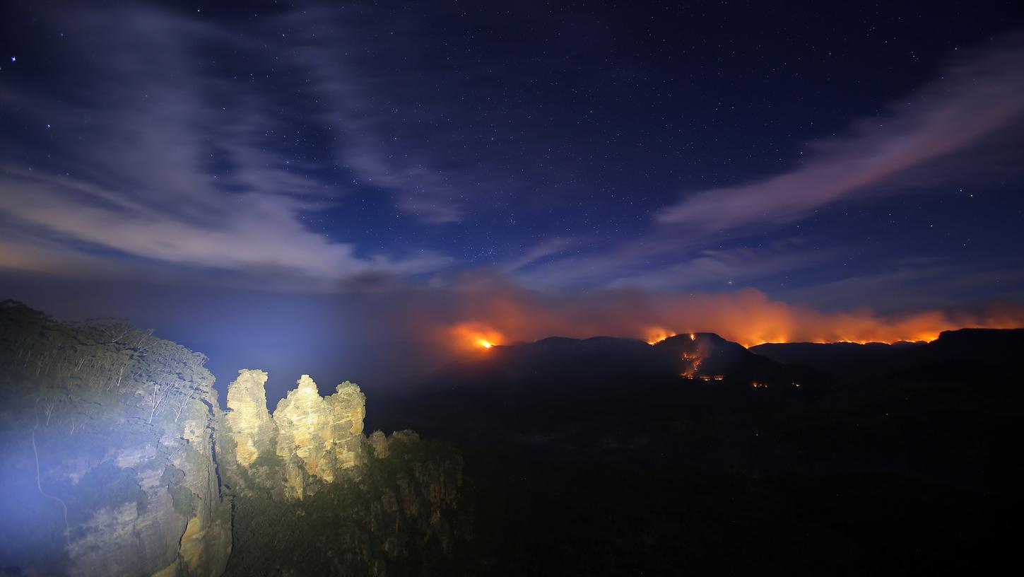 Eine Langzeitaufnahme zeigt die Felsformation Three Sisters, während im Hintergrund die Flammen des Green Wattle Creek Feuers zu sehen sind.