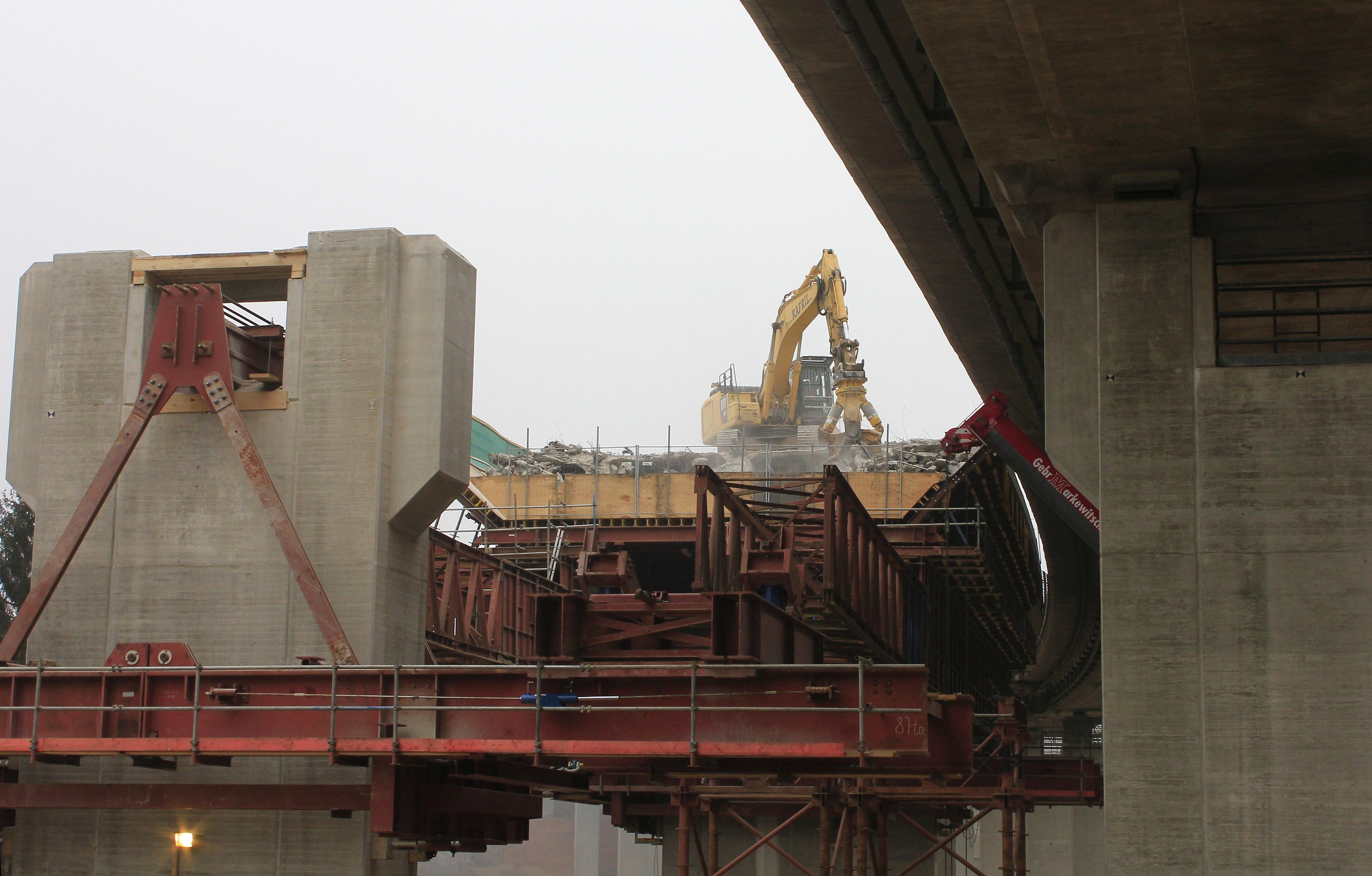 Ein Abrissbagger reißt, auf der ehemaligen Fahrbahn stehend, die Brücke nach und nach ein und lässt den Schutt in eine große Stahlwanne fallen.