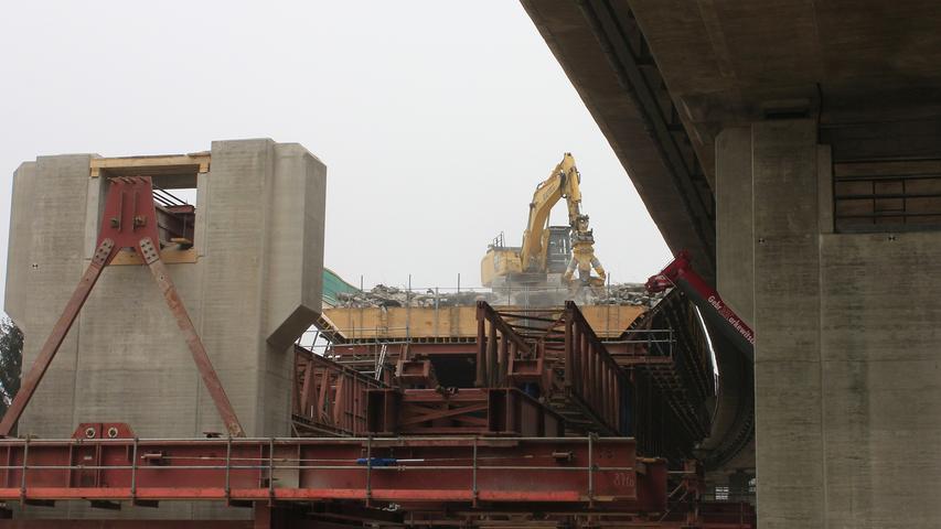 Ein Abrissbagger reißt, auf der ehemaligen Fahrbahn stehend, die Brücke nach und nach ein und lässt den Schutt in eine große Stahlwanne fallen.