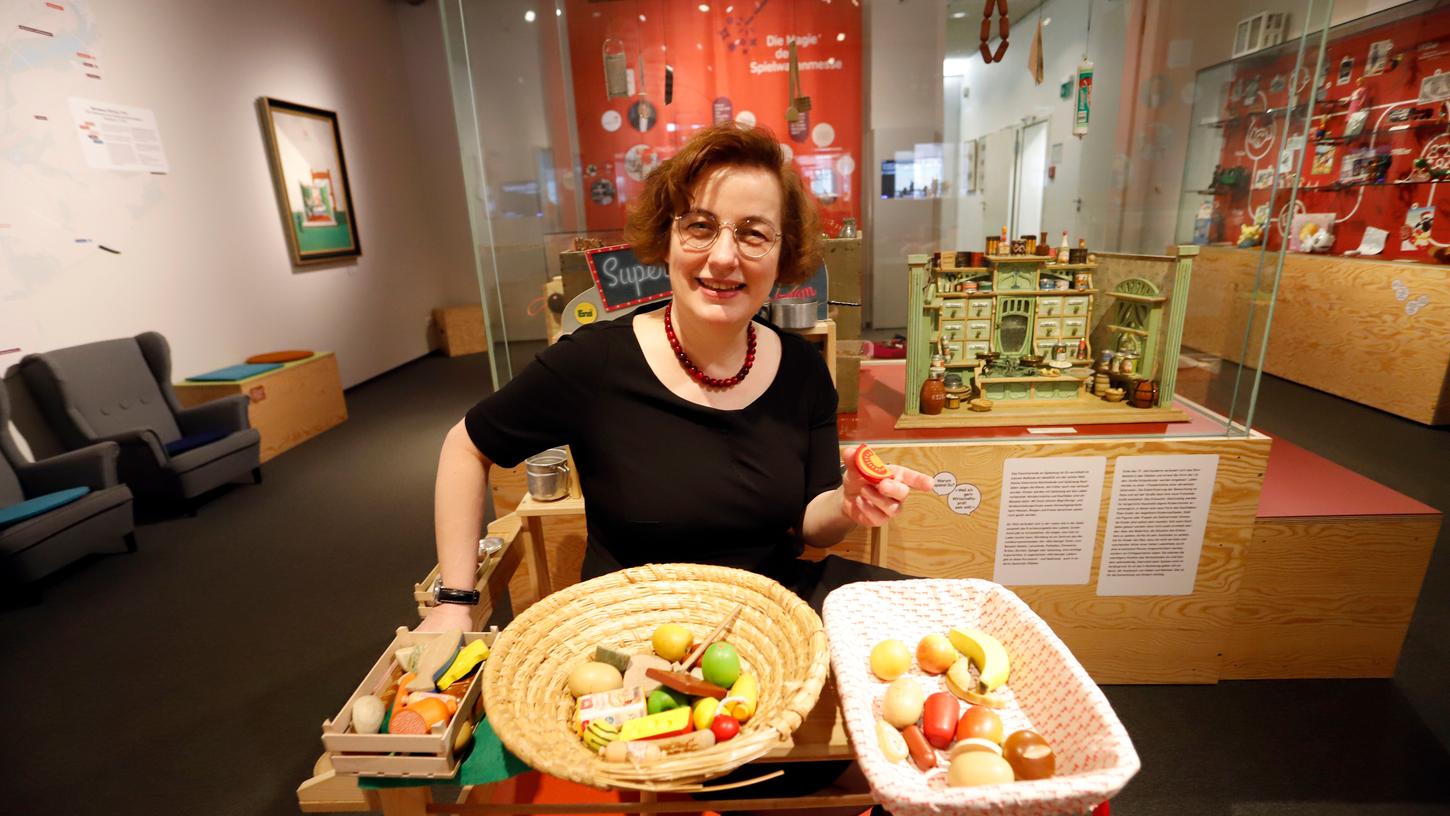 Karin Falkenberg, Leiterin des Spielzeugmuseums, sitzt  vor einer Vitrine mit  einem historischen Kaufmannsladen. Vor ihr liegen Holzfrüchte, mit denen die kleinen Besucher das Einkaufen nachspielen können.